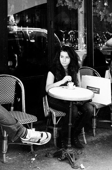 Black and white photo of a woman sitting alone at an outdoor café table, drinking from a cup. The table is small and round, situated between woven-patterned chairs. Reflections and bicycles are visible through the glass behind her. C'est le pied—a man's leg appears in the foreground.
