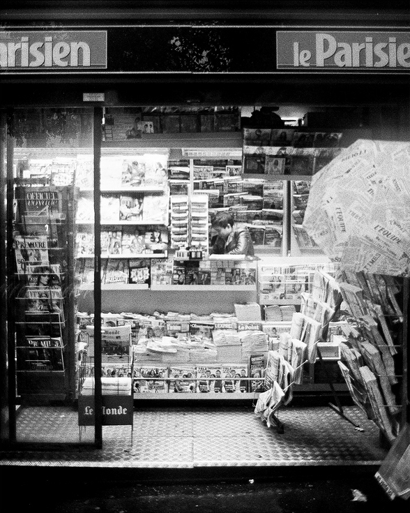 A black and white photo of a bustling kiosque at night. The stand is filled with magazines, newspapers, and various printed materials. The name "Le Parisien" is displayed at the top, and the front is adorned with stacks of publications.
