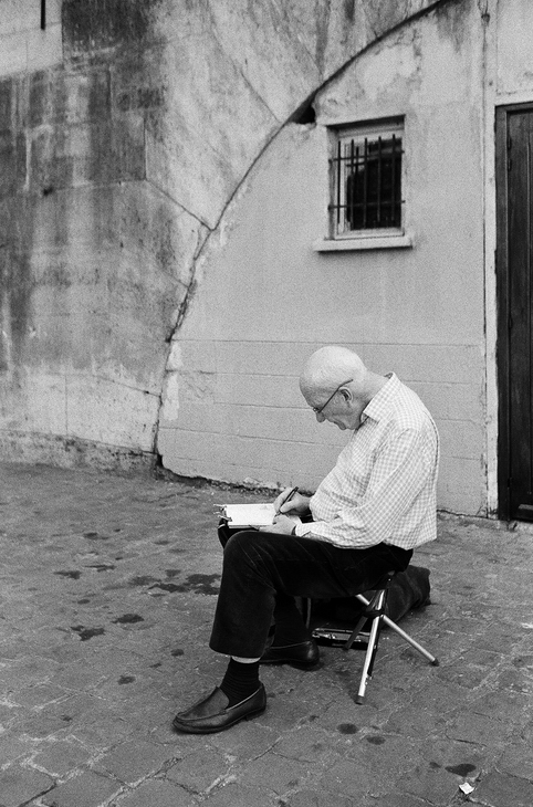 A bald man wearing glasses and a checkered shirt sits on a folding stool against a rough, partially graffitied wall. He is writing or drawing in his carnet de croquis placed on his lap, focused on his work in this candid black and white photograph.
