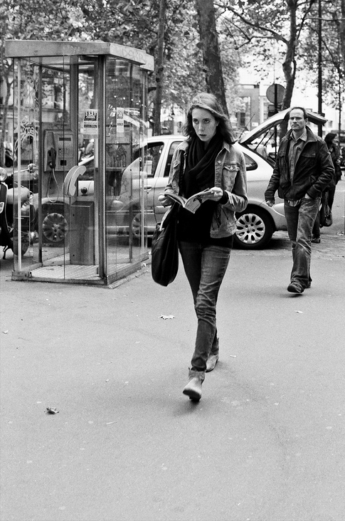 Black and white photo of Sophie Braque walking while reading a book on a city street. She is wearing a jacket, jeans, and a bag over her shoulder. Behind her, there is a man walking and a car near a telephone booth. Trees and more cars are visible in the background.
