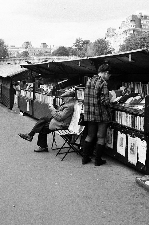 A black-and-white photo captures a street market bookstall. An elderly man sits and reads in front of one of the stalls, embodying the spirit of an amoureux des livres. Nearby, a person in a checkered coat and boots browses livres displayed on tables and shelves, with buildings forming the backdrop.