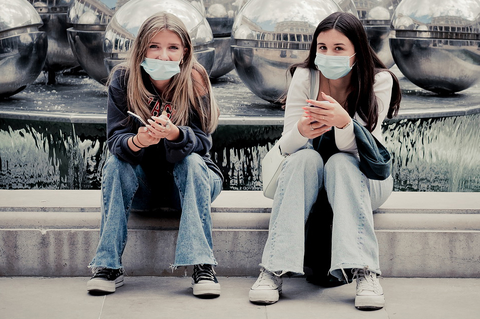 Two young women wearing face masks sit on a ledge in front of a modern fountain with metal spheres, each holding a smartphone and looking at the camera. Both wear jeans and casual tops.