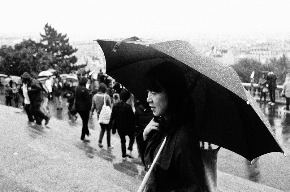 A woman holds an umbrella in the rain, walking past a crowd on wet steps. The photo is in black and white, with people in the background and trees to the left.