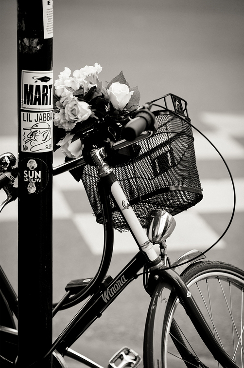 Dites le avec des fleurs A black bike with a front basket is parked against a pole adorned with various stickers. The basket contains a bunch of fleurs. The image is in black and white, adding a nostalgic, vintage feel.
