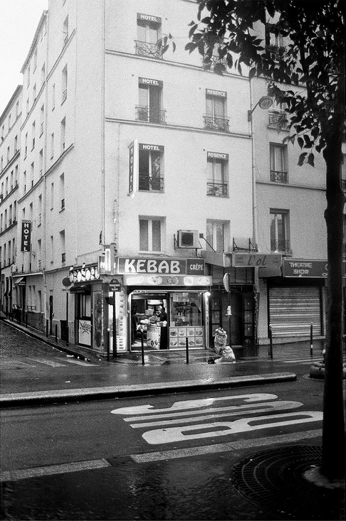 Black and white image of a street corner featuring a small kebab shop with a sign in French. The building above the shop has "HOTEL" signs. The urban scene shows wet ground, indicating recent rain. A hint of Mediterranean food lingers as grilled meat aromas mingle in the air near a partially visible tree on the right side.